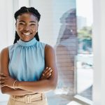 Ambitious young black businesswoman standing with her arms crossed by a window in an office alone