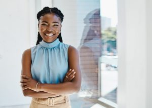 Ambitious young black businesswoman standing with her arms crossed by a window in an office alone