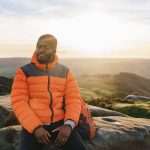 Man enjoying a peaceful moment while sitting on a rock at sunset in a scenic landscape with rolling hills and a colorful sky