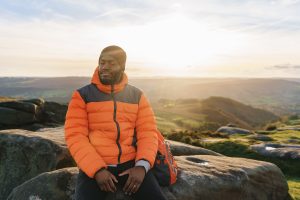 Man enjoying a peaceful moment while sitting on a rock at sunset in a scenic landscape with rolling hills and a colorful sky